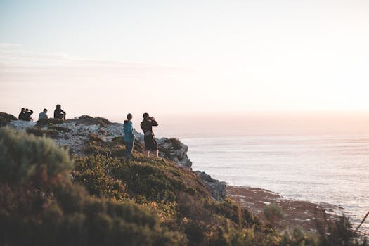 Full length travelers wearing casual clothes resting on rocky cliff and enjoying amazing scenery of calm rippling sea during amazing sunset