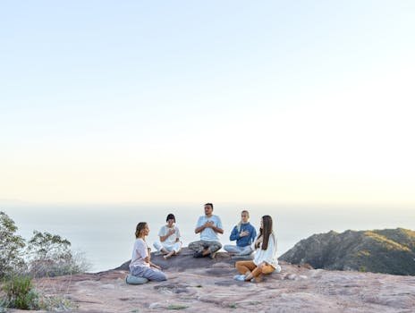 A diverse group meditates on a scenic mountaintop overlooking the sea, promoting relaxation and well-being.