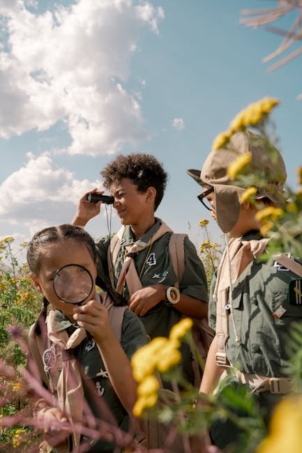 Three children in scout uniforms exploring nature with binoculars and magnifying glass on a sunny day.
